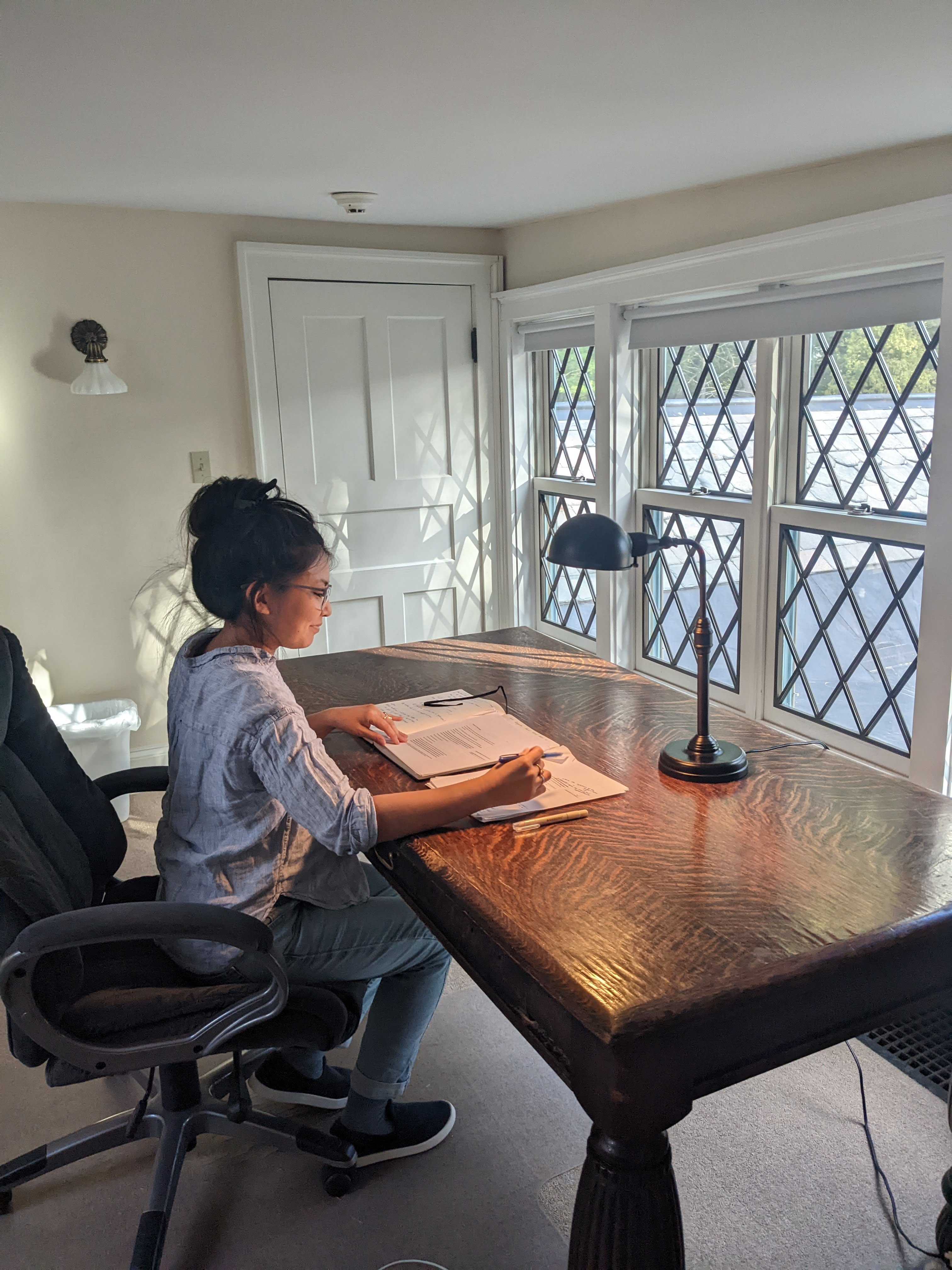 A person seated at a large wooden desk writing in a notebook, with a lamp and windows in the background.